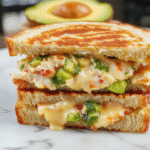 A vibrant grilled chicken avocado melt sandwich on a rustic wooden board. The sandwich features crispy toasted bread, gooey melted cheese, slices of ripe avocado, and tender grilled chicken. The sandwich is garnished with fresh greens and served with a side of salad, showcasing bright green, golden brown, and creamy textures.