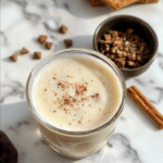 A steaming cup of vanilla cinnamon latte with swirls of cinnamon on top, placed on a rustic wooden table. The latte has a creamy foam with sprinkled cinnamon and vanilla bean pods beside the cup, styled with a cozy autumn backdrop including soft warm tones and subtle coffee shop ambiance.