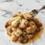 A close-up of a creamy mushroom chicken dish served on a white plate, featuring tender chicken breasts topped with a rich, velvety mushroom sauce, garnished with fresh herbs, set against a rustic wooden table with sautéed mushrooms and creamy sauce visible.